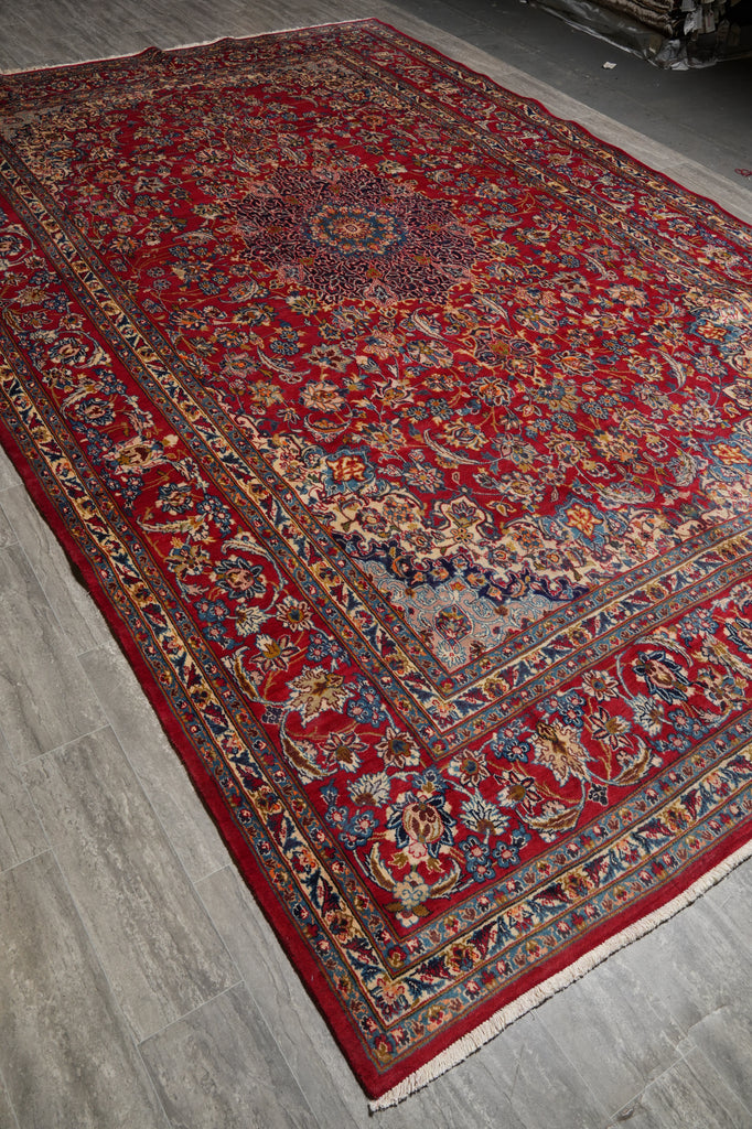 A traditional Persian-style area rug with a deep red field, intricate floral motifs, and a detailed border, laid out on a gray wood floor.