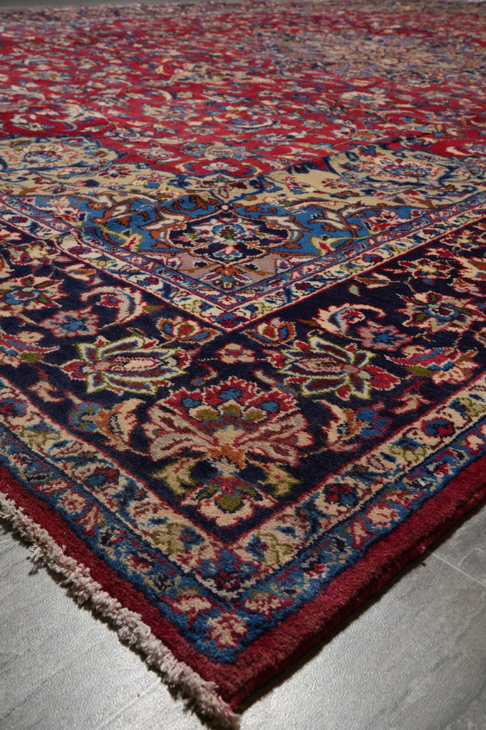 A traditional oriental area rug with an intricate floral pattern, shown in a close-up view highlighting its corner and fringe detail. The rug features a deep red field, a dark navy blue border, and accents of beige, blue, and green.
