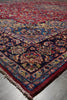 Load image into Gallery viewer, A traditional oriental area rug with an intricate floral pattern, shown in a close-up view highlighting its corner and fringe detail. The rug features a deep red field, a dark navy blue border, and accents of beige, blue, and green.