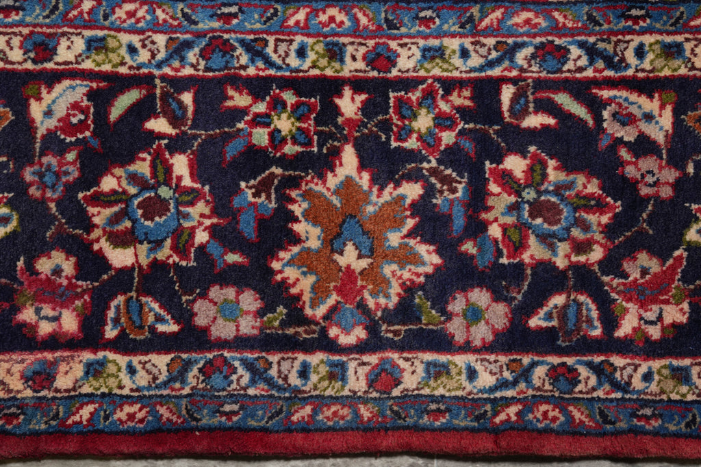 A close-up of a traditional patterned rug with a dark navy blue field, featuring intricate floral motifs in red, beige, blue, and green, bordered by detailed geometric and floral bands.