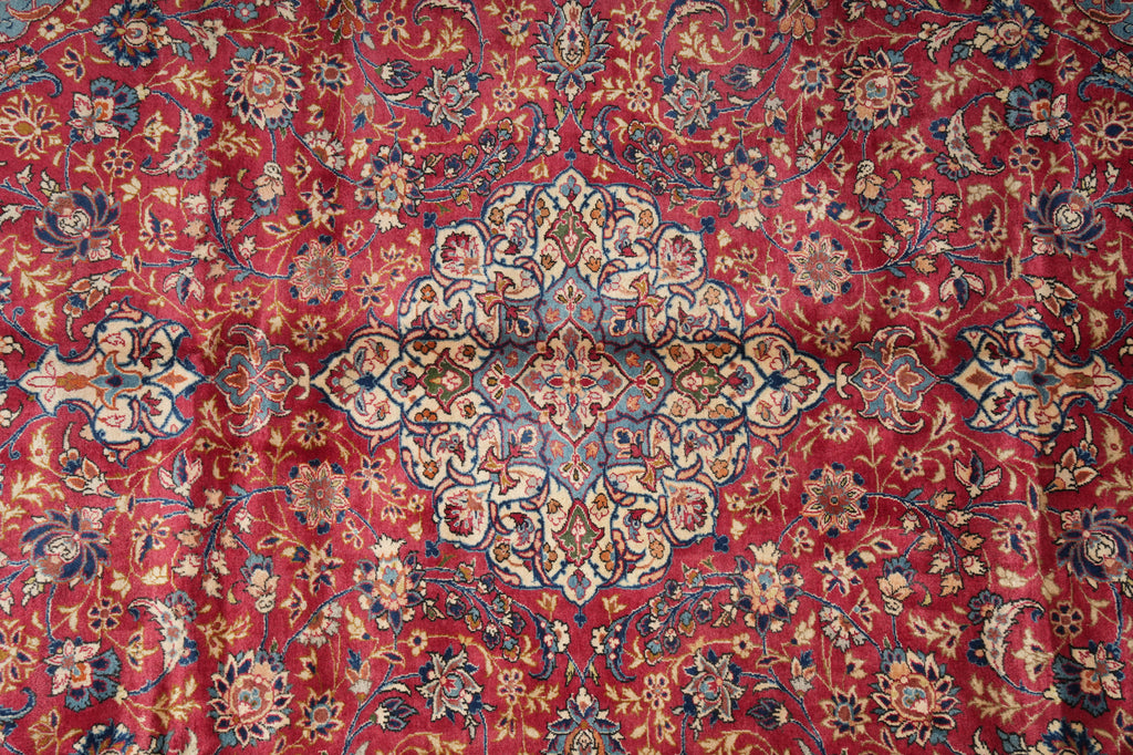 A close-up of an ornate Persian-style rug with a deep red background, featuring a large central medallion and intricate floral motifs in beige, blue, and gold.