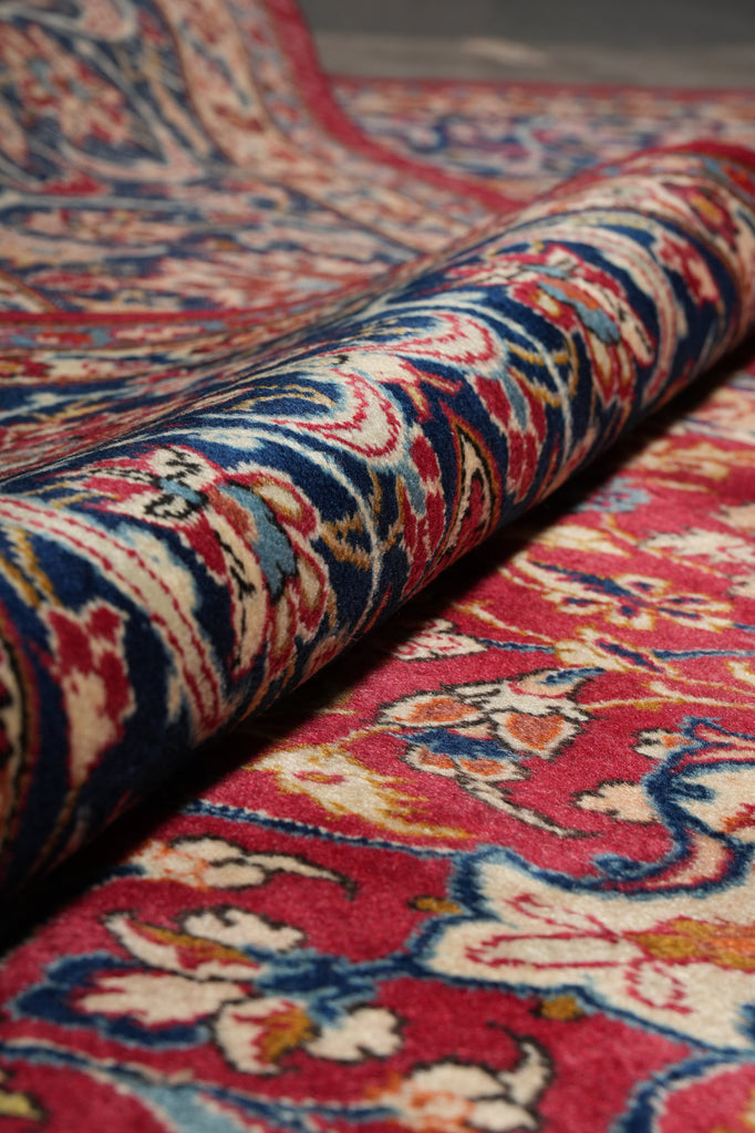 A close-up of a folded patterned rug showing a detailed floral and ornamental design in red, blue, cream, and beige tones.