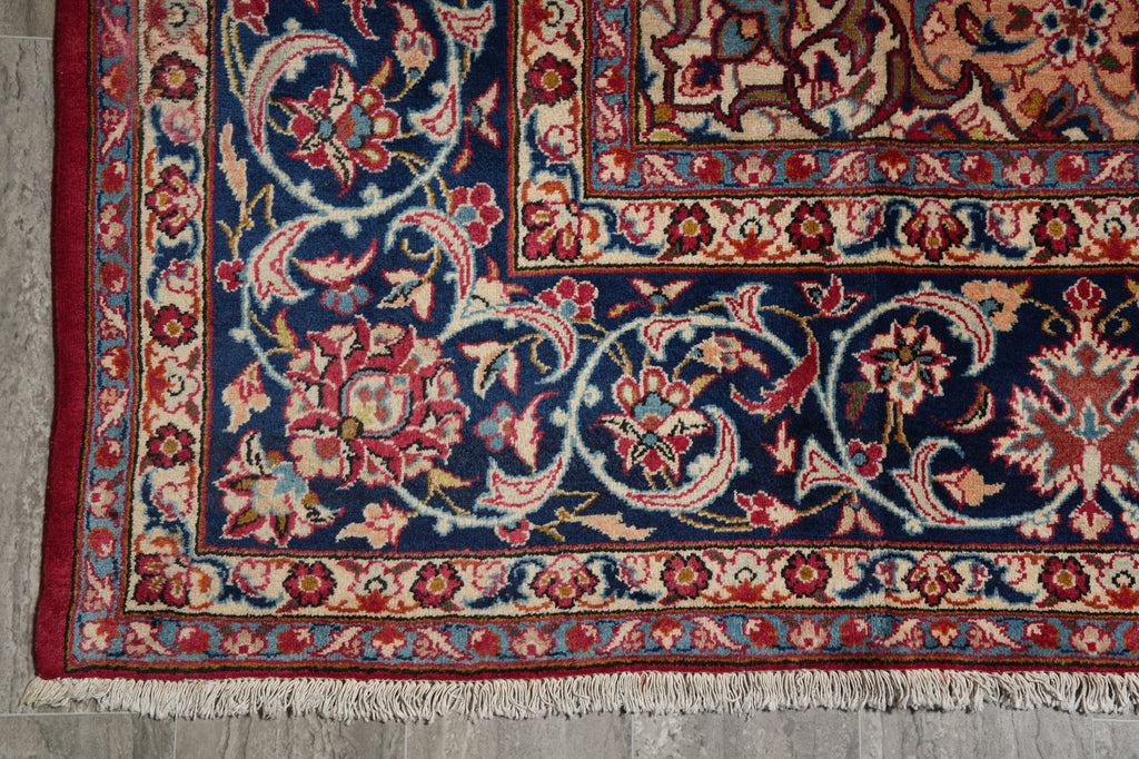 A close-up of an ornate Persian-style area rug corner, showing a deep navy blue field with intricate floral motifs in red, beige, and light blue, finished with white fringe.