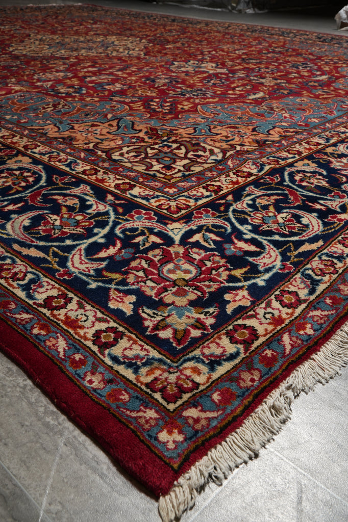 A traditional oriental-style area rug with an intricate floral pattern, featuring a deep red field, dark blue border, and cream fringe, shown laid out on a gray tiled floor.