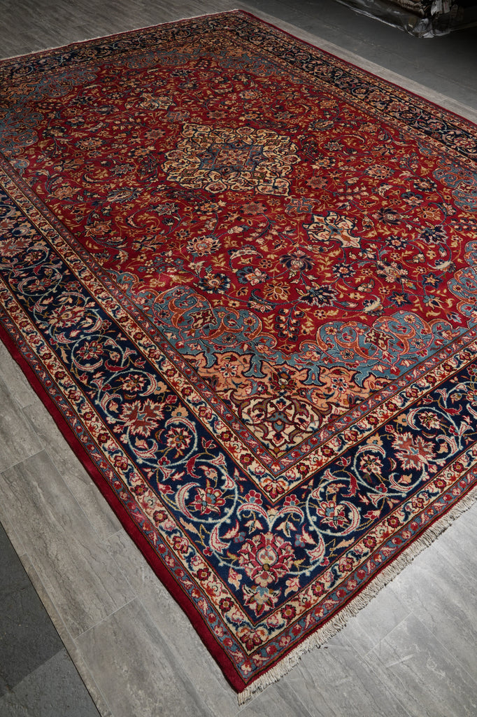 A traditional oriental area rug with a deep red field, ornate navy blue borders, and beige floral motifs, shown laid out on a gray wood floor.