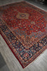 Load image into Gallery viewer, A traditional oriental area rug with a deep red field, ornate navy blue borders, and beige floral motifs, shown laid out on a gray wood floor.