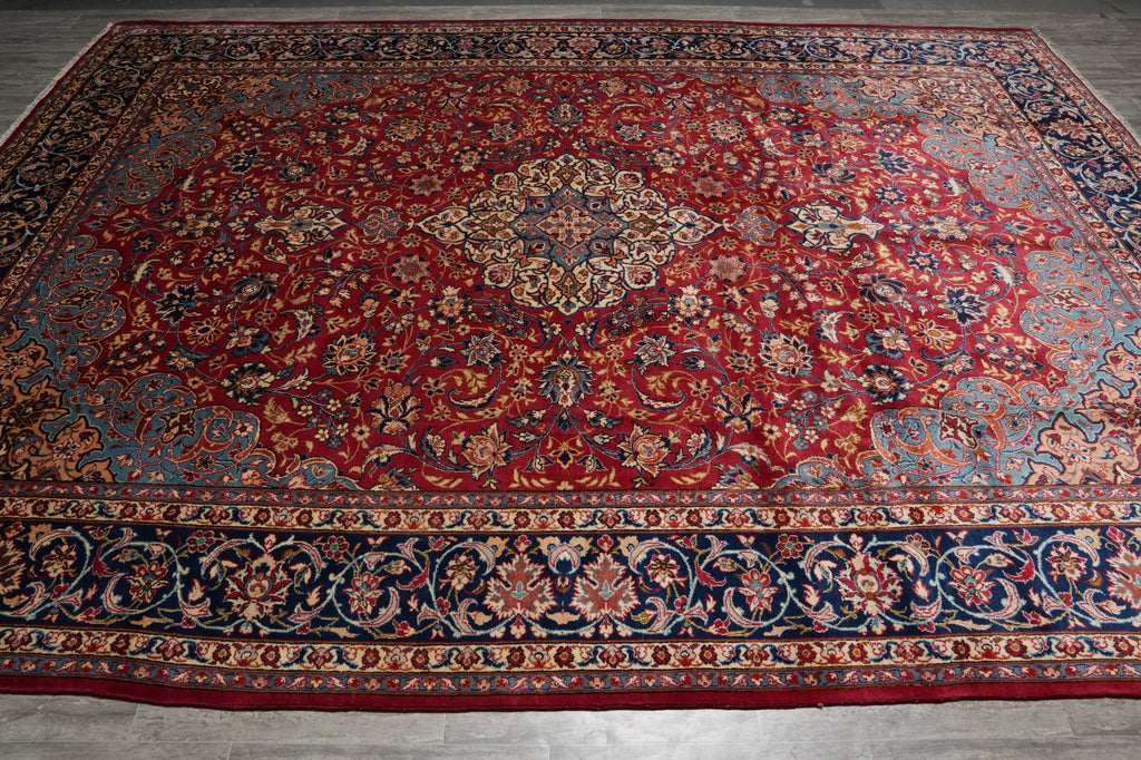 A traditional oriental area rug with a rich red field, central medallion, and detailed floral motifs in navy blue, beige, and gold, laid out on a gray floor.