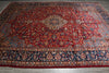 Load image into Gallery viewer, A traditional oriental area rug with a rich red field, central medallion, and detailed floral motifs in navy blue, beige, and gold, laid out on a gray floor.