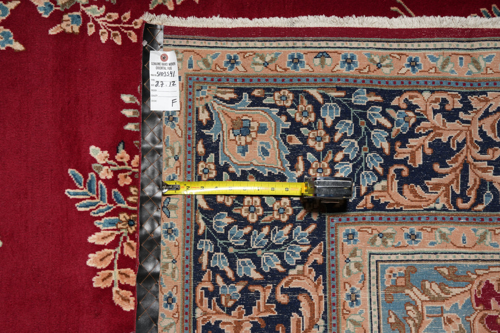 A close-up of an ornate patterned rug with a dark navy blue field, beige borders, and floral motifs in blue, green, and rust red. A yellow tape measure is placed horizontally across the rug to show its width, and a white product tag is visible on the left edge.