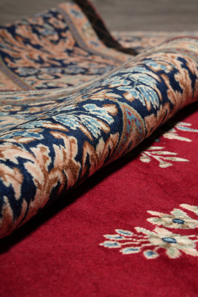 Close-up of a patterned rug with a rolled section showing a red field, cream floral motifs, and a central pattern in navy blue, tan, and light blue.