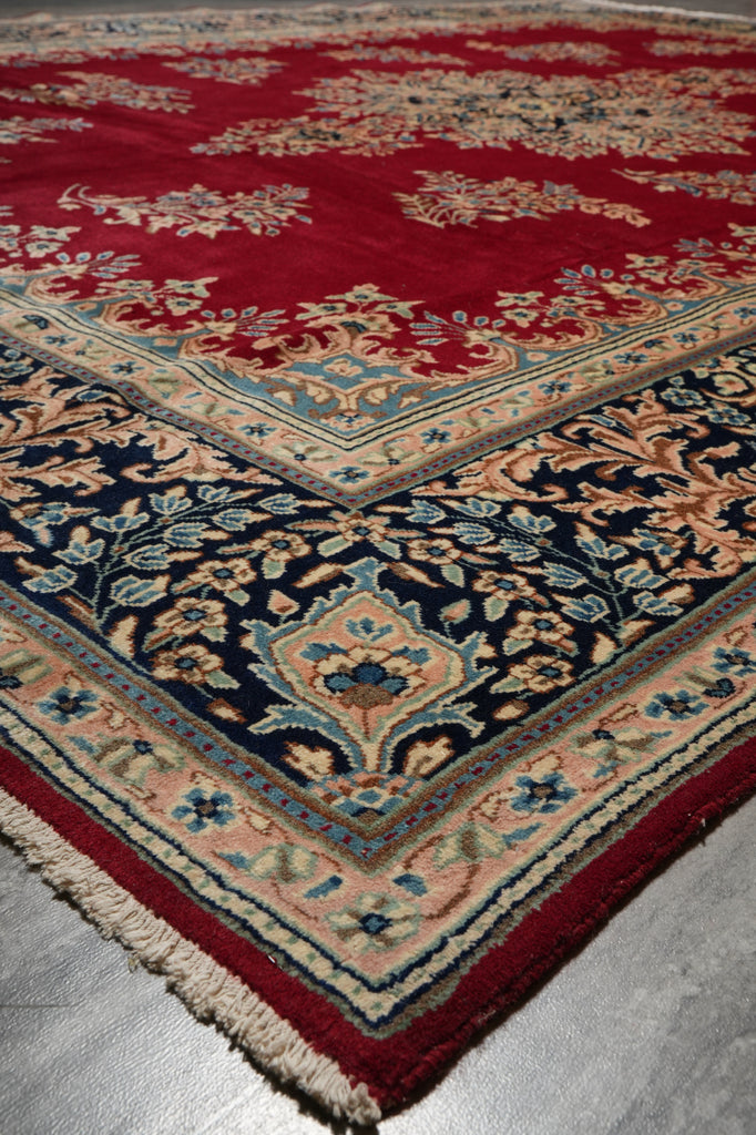 A close-up of a patterned area rug corner showing a deep red field, navy blue central section, peach and beige floral motifs, and a red border with fringe.