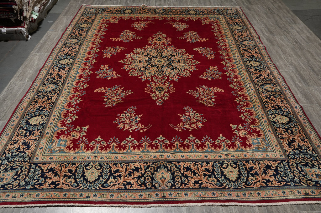 A traditional oriental area rug with a deep red field, featuring a central floral medallion and ornate borders in beige, navy blue, and green, laid out on a gray wooden floor.