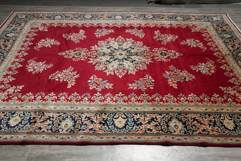 A traditional oriental area rug with a vibrant red field, featuring a central floral medallion and ornate borders in navy blue, beige, and gold, laid out on a gray tiled floor.