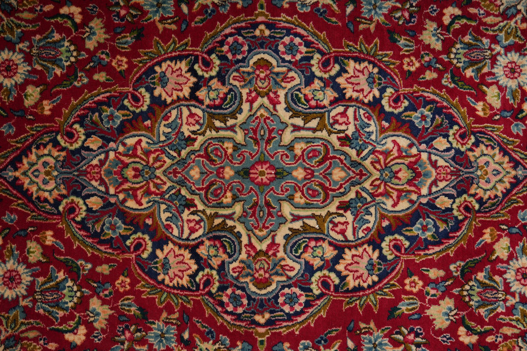 A close-up of an ornate traditional patterned rug with a deep red background and intricate floral motifs in blue, beige, green, and pink.