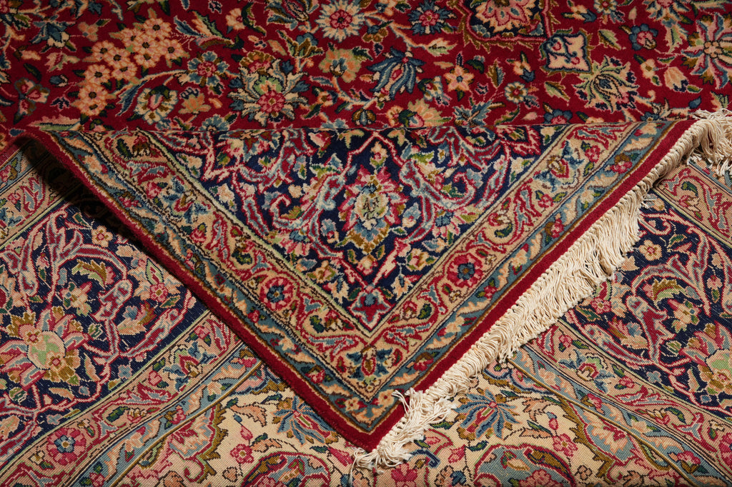A close-up of an ornate Persian-style area rug with a deep red field, intricate floral motifs in blue, beige, and green, and cream fringe along one corner.
