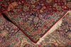 Load image into Gallery viewer, A close-up of an ornate Persian-style area rug with a deep red field, intricate floral motifs in blue, beige, and green, and cream fringe along one corner.