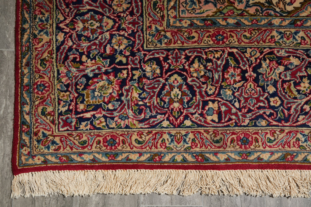 A close-up of an ornate traditional area rug with a complex floral and vine pattern in deep red, navy blue, beige, and green, featuring cream-colored fringe on the bottom edge.