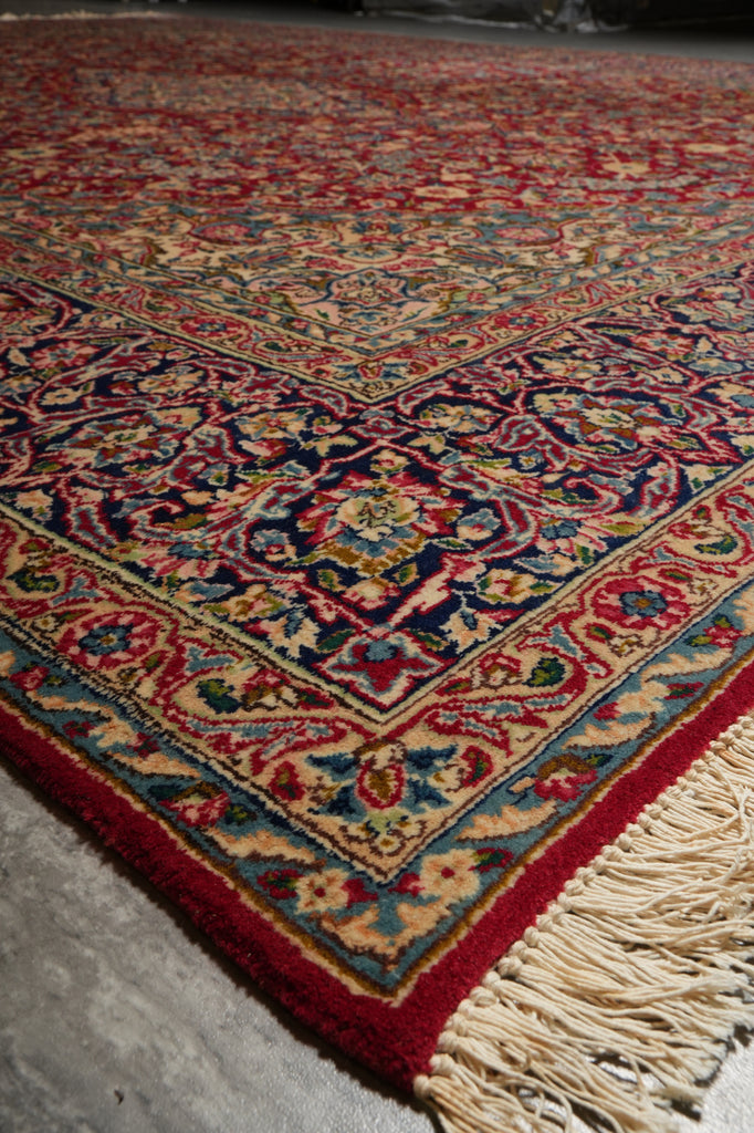 A close-up of a traditional patterned rug with cream fringe, featuring a red border and an intricate central design in red, blue, beige, and green tones.