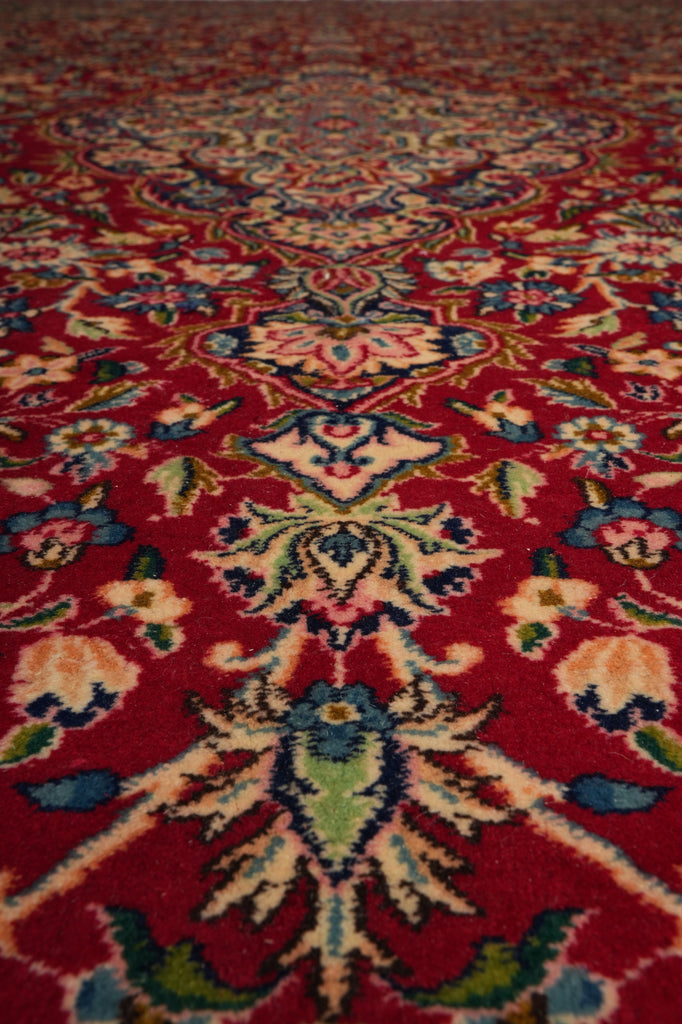 A close-up view of a traditional patterned rug with a deep red background and ornate floral motifs in cream, blue, green, and pink.