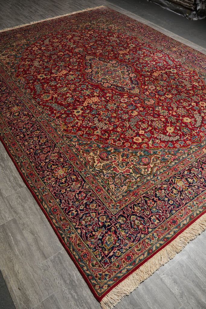 A traditional oriental area rug with a deep red field, intricate floral motifs, and a detailed border, shown laid out on a gray wood-look floor.