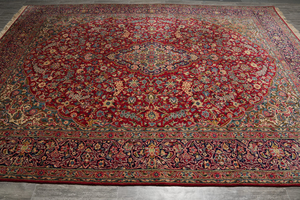 A large traditional Persian-style area rug with a dominant deep red field, featuring an intricate central medallion and detailed floral motifs in blue, green, beige, and gold, laid out on a gray tiled floor.