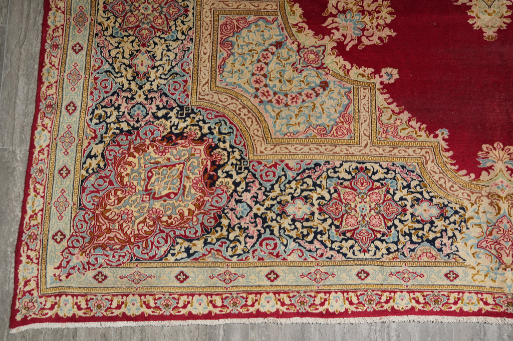 A close-up of an ornate traditional area rug showing its corner detail, featuring a deep red field, a navy blue central field, and cream, gold, and pink floral patterns.