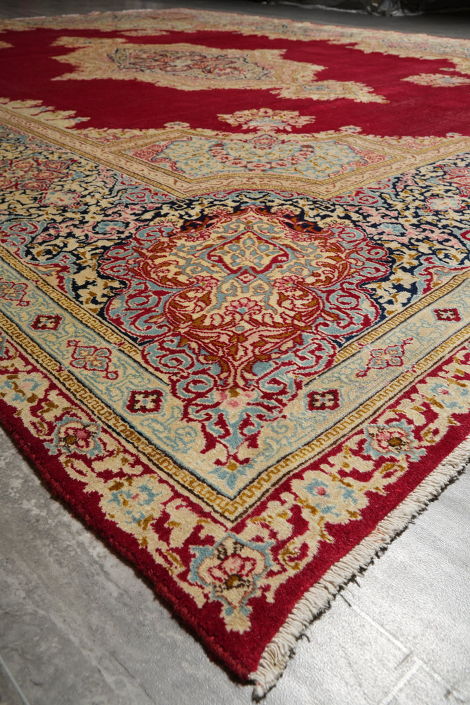 A close-up of a traditional oriental rug with a deep red field, cream and beige borders, and a central medallion in red, blue, and beige, laid on a gray floor.