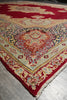 Load image into Gallery viewer, A close-up of a traditional oriental rug with a deep red field, cream and beige borders, and a central medallion in red, blue, and beige, laid on a gray floor.