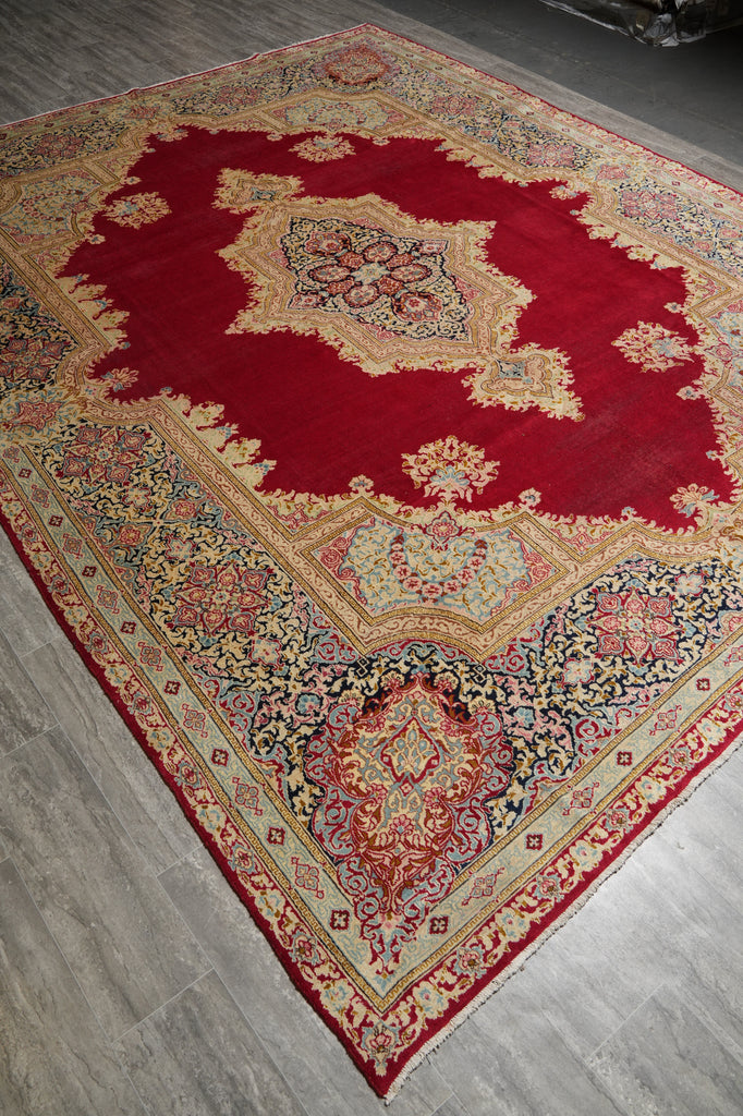 A traditional oriental area rug with a deep red central field, beige and gold ornamental medallions, and a multicolored floral border, shown laid out on a gray wood floor.