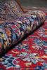 Load image into Gallery viewer, A close-up of a rolled section of a traditional patterned rug resting on a matching flat section of the same rug. The rug features an intricate floral and geometric design in red, navy blue, beige, and orange tones.