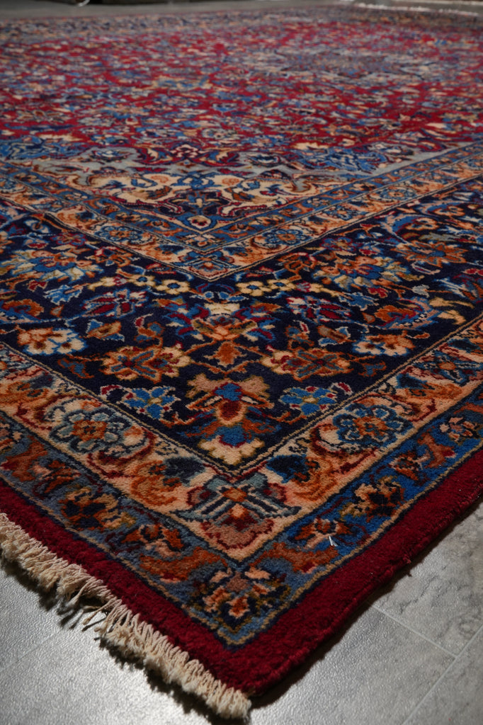 A close-up of a traditional oriental rug showing its corner, featuring a deep red border, cream fringe, and an intricate floral pattern in navy blue, rust orange, and beige on a gray floor.
