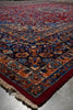 Load image into Gallery viewer, A close-up of a traditional oriental rug showing its corner, featuring a deep red border, cream fringe, and an intricate floral pattern in navy blue, rust orange, and beige on a gray floor.