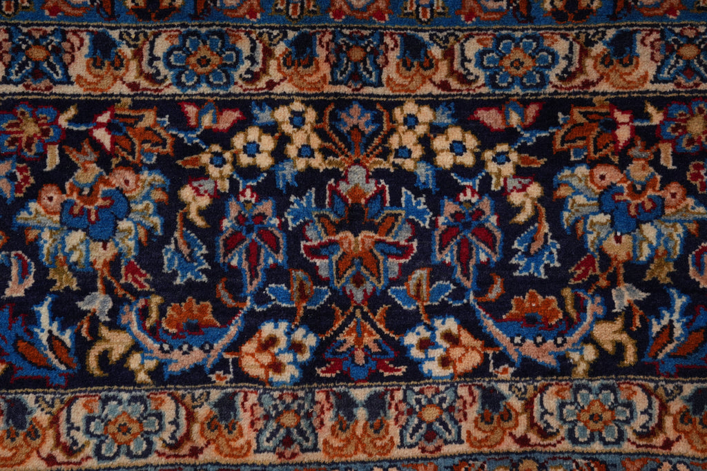 A close-up of an ornate patterned rug with a dark navy blue field, featuring intricate floral motifs in blue, rust red, beige, and cream, framed by decorative borders.