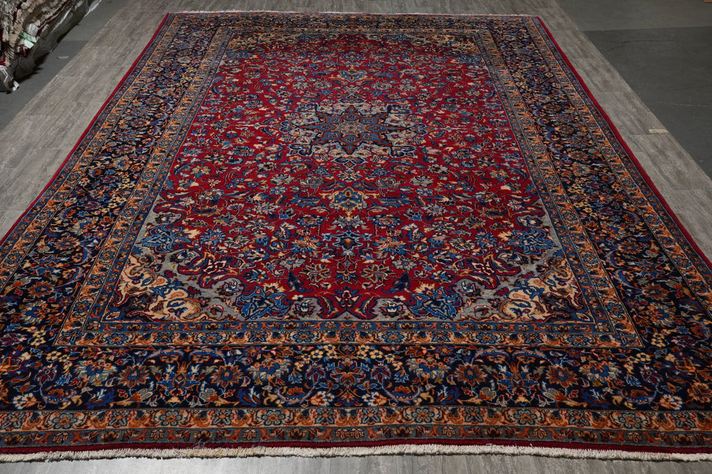 A traditional Persian-style area rug with a deep red central field, detailed blue and beige floral motifs, and an ornate multi-colored border, shown laid out on a gray floor.