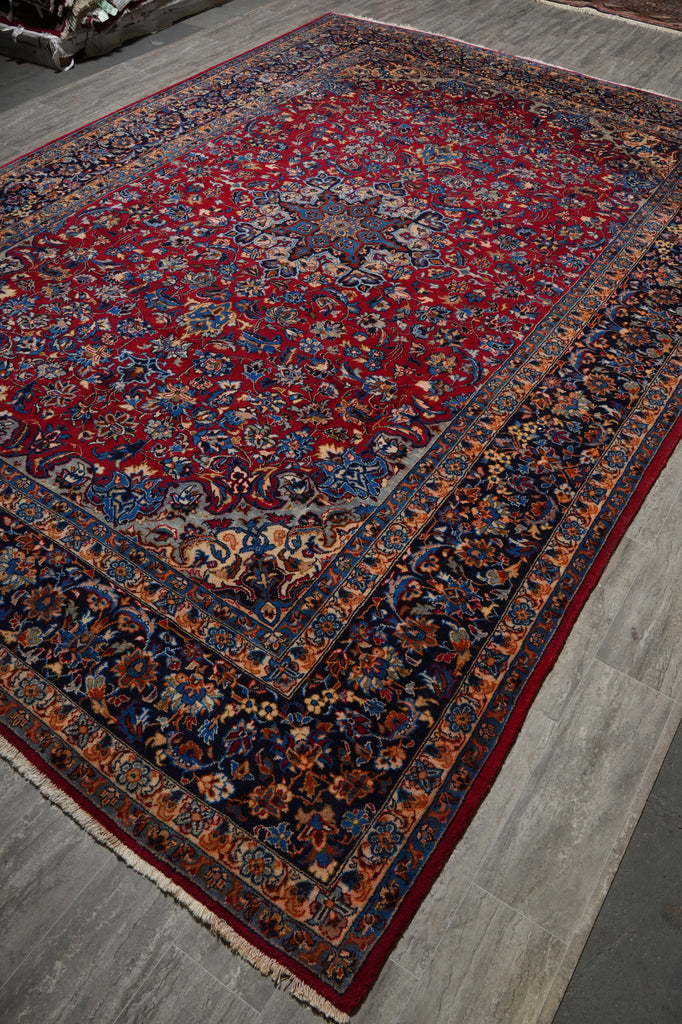 A traditional Persian-style area rug with a red field, dark navy borders, and intricate floral motifs in blue, gold, and beige.