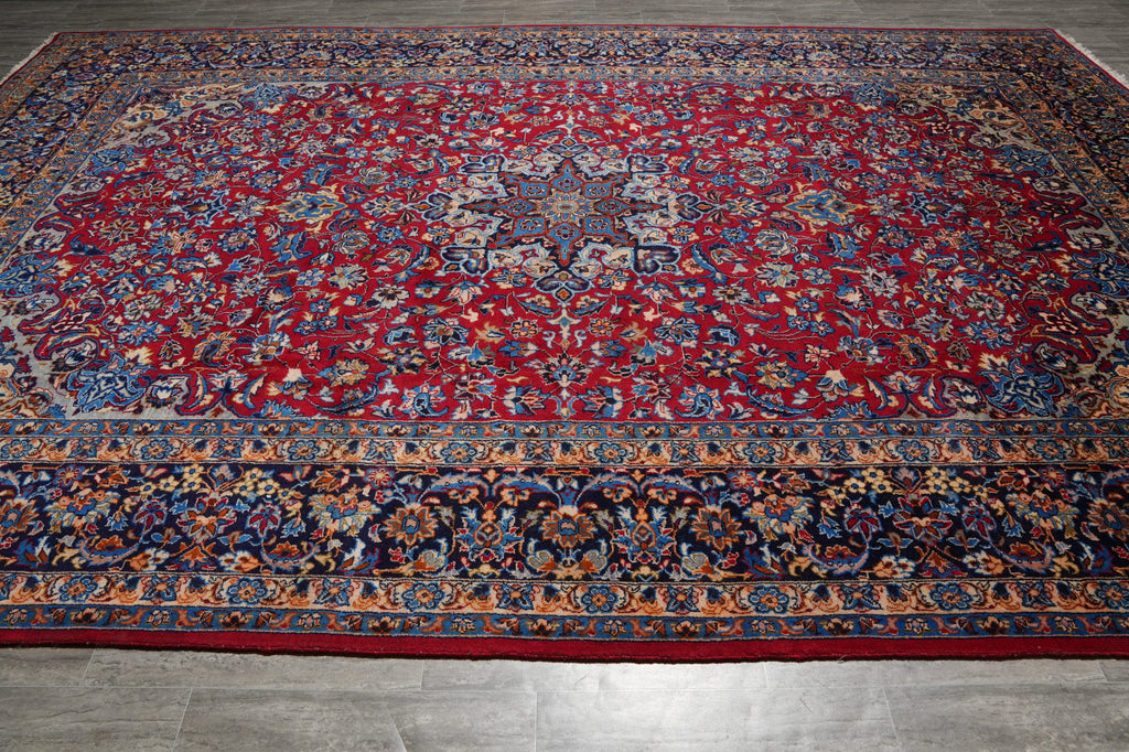 A traditional oriental area rug with a deep red field, intricate floral motifs, and detailed blue, beige, and gold borders, shown laid out on a gray tiled floor.