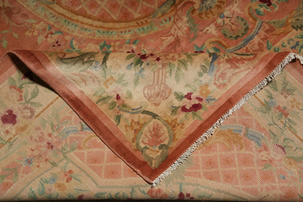 A close-up of a folded vintage patterned textile, likely a rug or tapestry, featuring a warm peach, pink, and beige floral design with fringed edges.