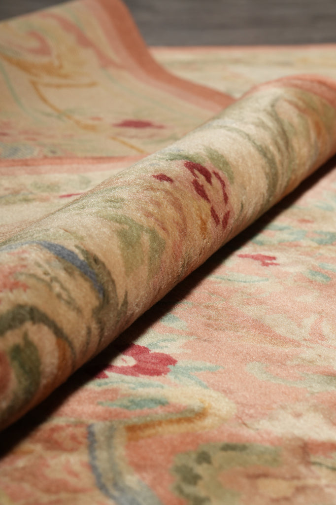 A close-up of a rolled area rug showing its detailed pattern and texture. The rug features a soft, warm beige base with a traditional floral motif in muted shades of pink, sage green, and red.