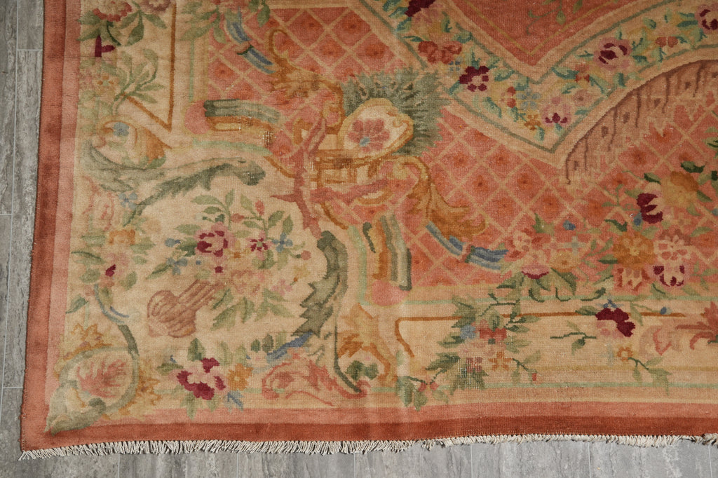 A close-up of an ornate traditional rug corner, showing a peach and beige field with floral motifs, green foliage, and a peach border, laid on a gray floor.