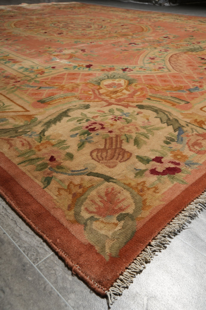 A close-up of an ornate area rug with a peach and rust border, cream field, and multicolored floral and scroll motifs, shown on a gray tiled floor.