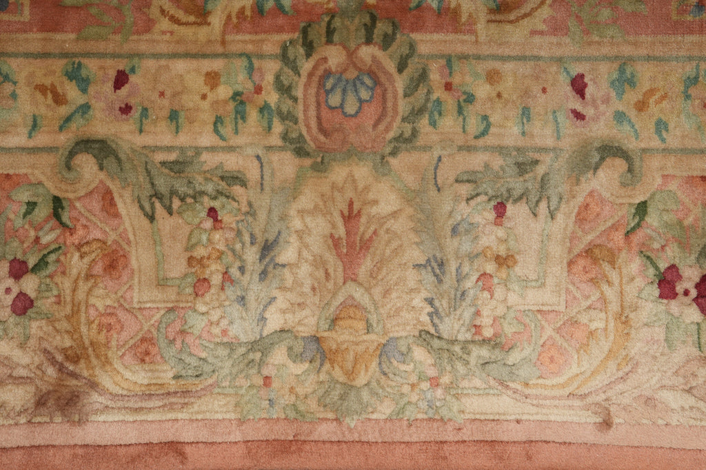 A close-up of an ornate vintage-style rug featuring a central floral medallion and scrolling motifs in muted pink, beige, green, and blue tones.