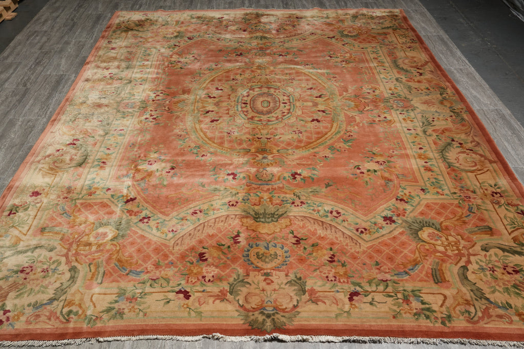 A traditional oriental area rug with a faded coral-pink center, beige borders, and multicolored floral and medallion motifs, laid out on a gray floor.