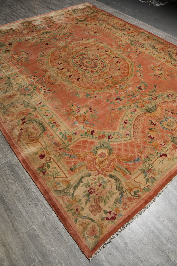 An ornate rectangular area rug with a warm terracotta and beige base, featuring intricate floral and medallion patterns in muted green, red, and blue, laid out on a gray wood floor.
