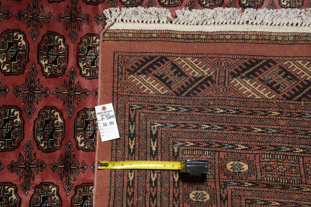 A close-up of a patterned area rug with a red field, dark navy geometric border, and cream fringe, shown alongside another red patterned rug and a yellow tape measure for scale.