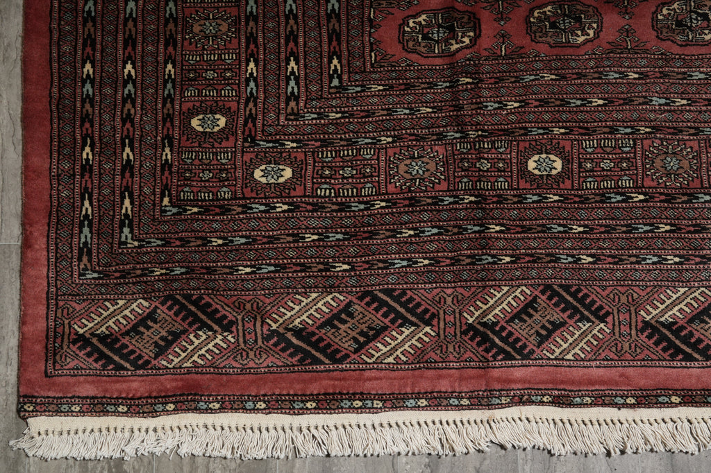 A close-up of a traditional patterned rug corner, showing a deep red field with black, beige, and cream geometric borders and white fringe tassels along the bottom edge.