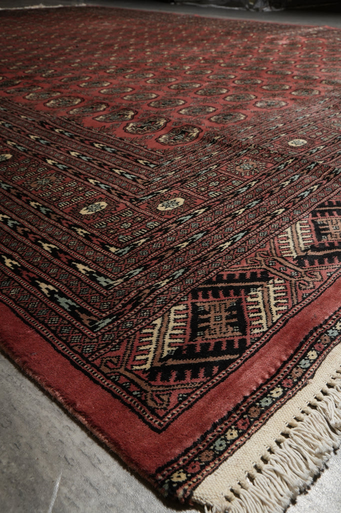 A close-up of a patterned area rug with a deep red field, dark navy borders, and cream fringe, photographed on a gray floor.