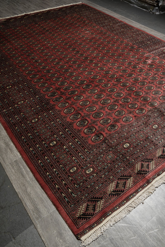A traditional Persian-style area rug with a deep red field, intricate black and beige geometric patterns, and a fringed edge, shown laid out on a gray tiled floor.