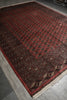 Load image into Gallery viewer, A traditional Persian-style area rug with a deep red field, intricate black and beige geometric patterns, and a fringed edge, shown laid out on a gray tiled floor.