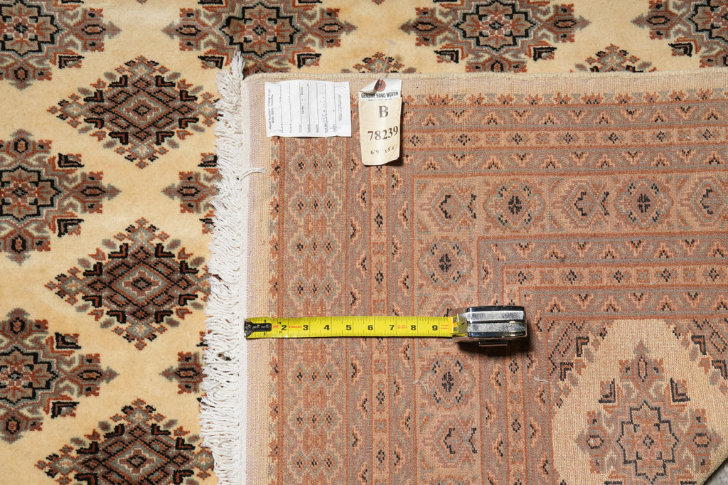 A patterned area rug shown folded with a tape measure laid across it to indicate size. The rug has a beige, brown, and rust-orange geometric design with white fringe.