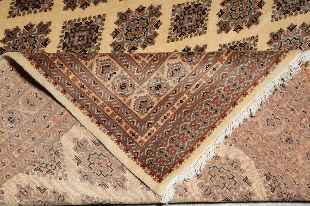 A close-up of a patterned area rug with a triangular section folded over to show the underside and fringe. The rug features intricate geometric and floral motifs in beige, brown, and muted red tones.